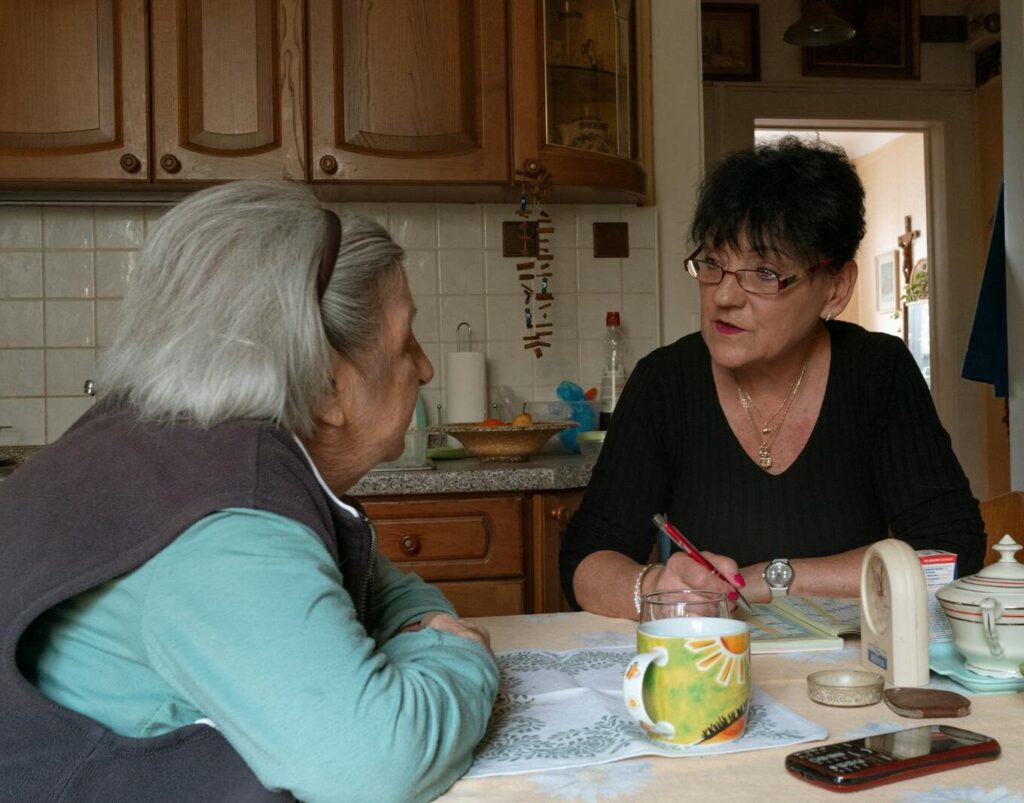 Senior woman and caregiver having a conversation in a cozy Prague kitchen setting.